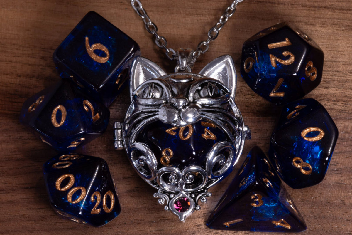 Cat pendant in a silver finish with a pink crystal, holding a dark blue D20, beside a dark blue dice set with gold numbers on wood